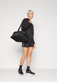Saco de ginásio preto com detalhes em malha, formato retangular e alça ajustável. Modelo está a usar roupa atlética preta e ténis. O fundo é simples.