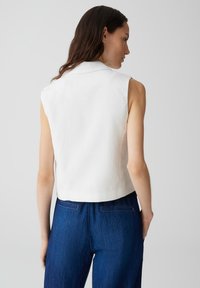 Top corto bianco senza maniche con colletto a punta e texture liscia, abbinato a jeans blu scuro a vita alta con tasche posteriori.