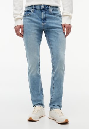 Jean slim bleu clair porté avec des baskets blanches et un haut blanc à manches longues, montré des hanches jusqu'aux pieds.