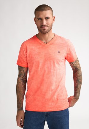 V-hals T-shirt in fel koraal met een gestructureerde afwerking, korte mouwen en een klein logodetail op de borst. Gecombineerd met spijkerbroeken.