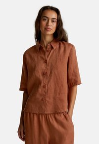 Donna che indossa una camicia botton-up di lino color ruggine a maniche corte e pantaloni alti coordinati, in piedi con una mano in tasca.