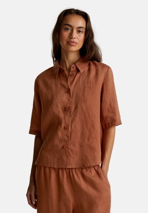 Vrouw in een roestkleurig linnen overhemd met knopen en korte mouwen, en bijpassende hoge taille broek, staand met één hand in de zak.