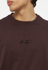 Κοντινή λήψη ενός ατόμου που φορά σκούρο καφέ μπλουζάκι με το κέντημα "Your Story" μαύρο στο στήθος και ασημένιο κολιέ αλυσίδα.