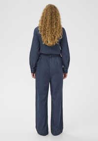 Ženska z dolgimi kodrastimi lasmi, oblečena v temno modro denim kombinezon s pasom, stoji obrnjena stran od enostavne bele ozadje.