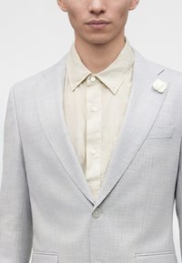 Homme portant une veste de costume gris clair avec une broche fleur blanche et une chemise beige boutonnée sur un fond uni.