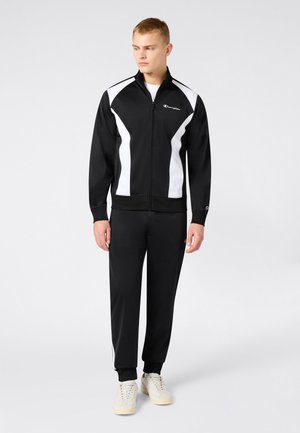 Młody mężczyzna w czarno-białej sportowej kurtce na zamek i pasujących spodniach, w białych trampkach, stoi na jasnym, jednolitym tle.