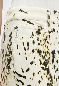Gros plan d’un pantalon couleur crème avec un motif abstrait éclaboussé noir et jaune et une étiquette de marque Modstrøm visible près de la poche avant.