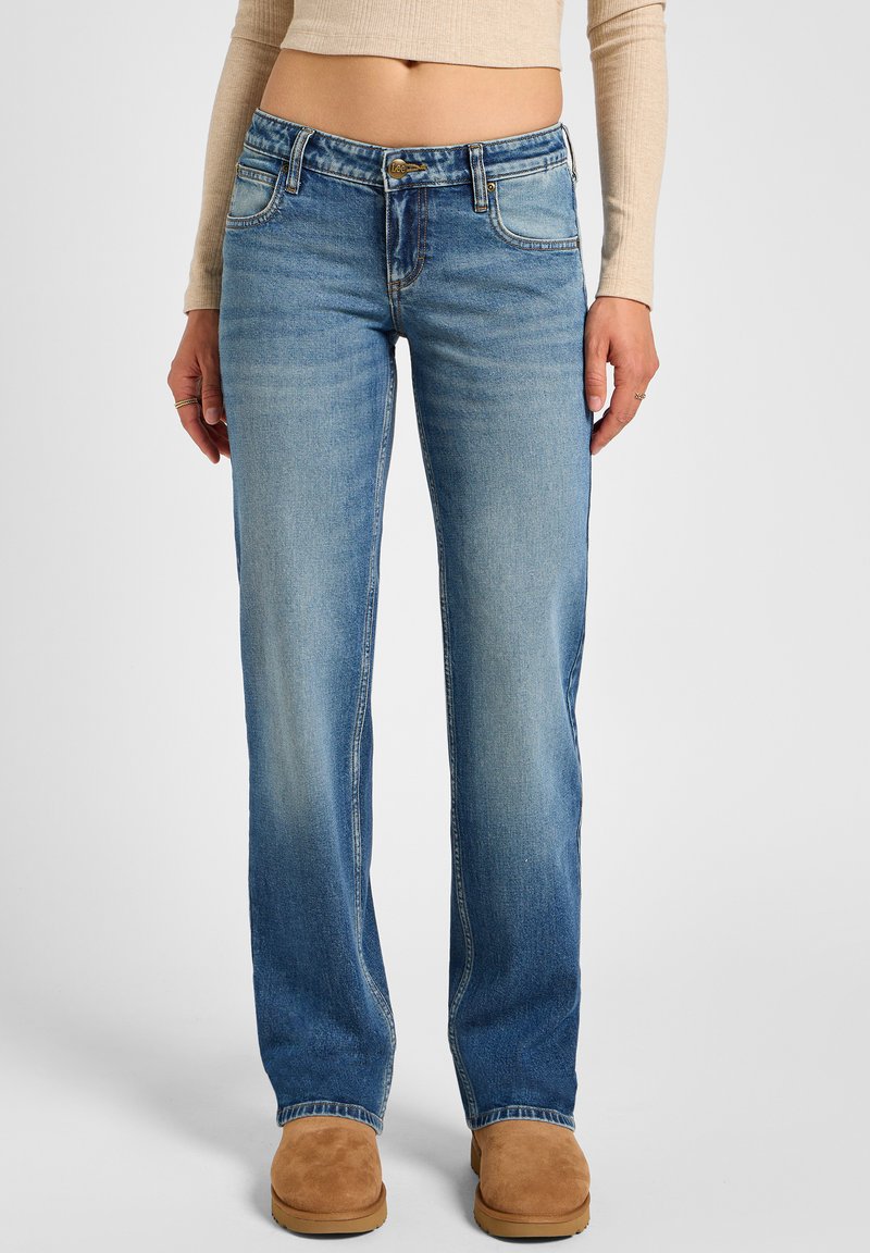 Los jeans de pierna recta en azul claro presentan una cintura de media altura, un ligero desgaste y un diseño clásico de cinco bolsillos. Combinados con botas de ante en color beige.