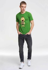 Green cotton t-shirt featuring a cartoon bee design with yellow body and black stripes. Paired with dark gray jeans and white sneakers.