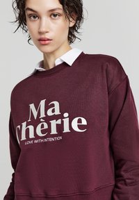 Giovane con capelli corti e bagnati che indossa una felpa bordeaux sopra una camicia bianca con colletto, con il testo "Ma chérie LOVE WITH INTENTION".