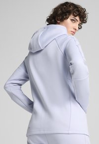 Haut de survêtement lilas clair avec un design ajusté, en tissu doux et avec un cordon de serrage. Présente des détails de panneaux d'épaule et latéraux sans couture. Texture lisse.