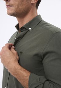 Chemise boutonnée vert foncé en tissu texturé, avec un petit accent brodé et des boutons en argent sur le col.