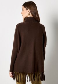 Brązowy, oversize sweter z prążkowanym wysokim kołnierzem, długimi rękawami i frędzlami na dole. Miękka textura i luźny krój.