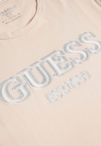 Camiseta de algodón beige con cuello redondo y logo plateado en relieve "GUESS EST. 1981" en el frente, textura suave y diseño ajustado.