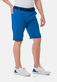Blaue Shorts aus einem Baumwollmischgewebe mit umgeschlagenen Säumen, seitlichen Taschen und kräftigen roten Nähakzenten. Tragen mit weißen Sneakers.