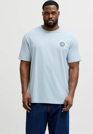 Man met kort haar en baard, gekleed in een lichtblauw T-shirt met een klein rond logo op de borst en donkerblauwe spijkerbroek, staand tegen een effen achtergrond.