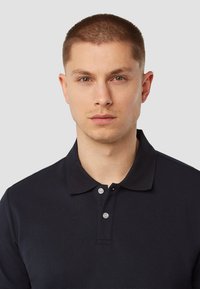 Marineblauwe poloshirt met een klassieke kraag, twee grijze knopen en korte mouwen. Het materiaal lijkt textuur te hebben en is ademend.
