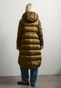 Lungo cappotto imbottito verde oliva con cappuccio, caratterizzato da un design trapuntato e polsini elastici, indossato sopra jeans blu e scarpe marroni.