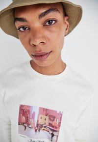 Sweatshirt blanc avec une impression graphique d'une scène de rue à New York, présentant un chapeau seau beige et une texture de peau lisse.