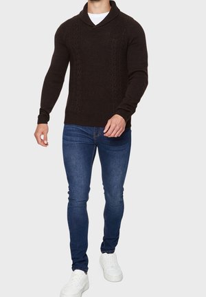 Homme portant un pull en laine noir, un jean slim bleu, des baskets blanches, marchant devant un fond clair uni.