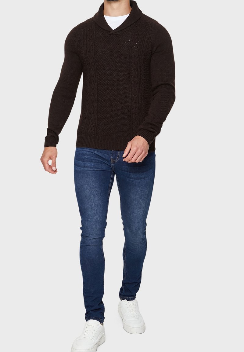 Homme portant un pull en laine noir, un jean slim bleu, des baskets blanches, marchant devant un fond clair uni.