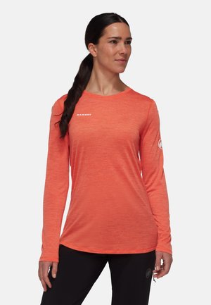 Kobieta w pomarańczowej długiej koszulce Mammut i czarnych spodniach, stoi z prawą ręką zrelaksowaną i patrzy na bok.