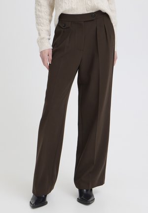 Femme portant un pantalon large marron foncé avec des boutons à la taille, un pull en maille torsadée crème et des bottines noires.