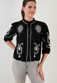 Schwarze knopflosse Bluse mit weißen gestickten Blumenmustern, ausgestattet mit einem Stehkragen und puffierten Ärmeln. Baumwollstoff mit strukturiertem Finish.