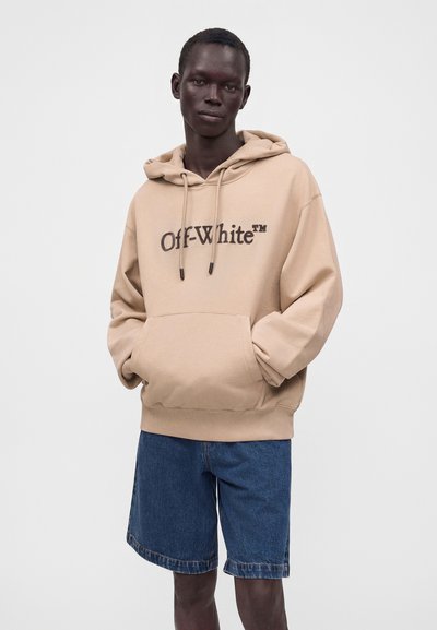 Jeune homme portant un sweat à capuche beige Off-White avec des poches et un short en jean bleu longueur genou, debout devant un fond clair uni.
