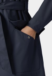 Hand in de zak van een donkerblauwe waterdichte jas met klittenbandsluiting en doorgestikte naden, afgebeeld tegen een effen lichte achtergrond.