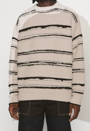 Pull beige à rayures horizontales noires irrégulières porté par une personne avec un pantalon foncé présentant des coutures blanches et de grandes poches avant.