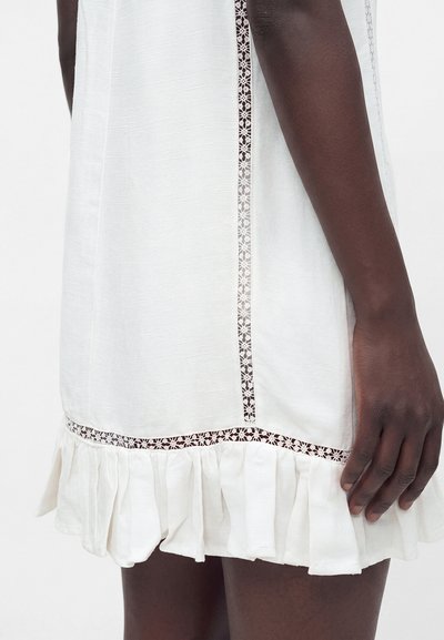 Bras à la peau foncée reposant à côté d'une robe blanche avec une bordure en dentelle florale et un ourlet froncé à hauteur de mi-cuisse sur un fond clair.