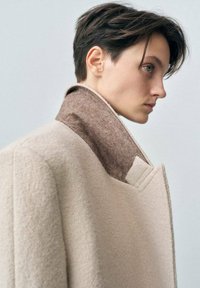 Cappotto in misto lana di un beige chiaro con colletto in feltro marrone. Presenta un design pulito e strutturato con tasche laterali nascoste.