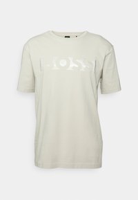 Světle béžové tričko z bavlny s krátkými rukávy, s reflexním logem "BOSS" přes hrudník. Hladká textura a volný střih.