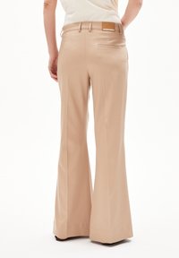 Pantalons évasés beiges avec une texture lisse, présentant une poche arrière et une étiquette marron sur la taille. Conception devant plat et coupe ajustée.