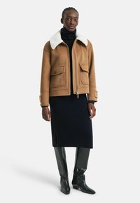 Brown wool jacket with white faux fur collar, two front pockets, zip closure, worn over a navy sweater and knee-length skirt; black leather boots.