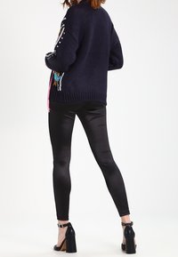 Námořnicky modrý pletený oversized kardigan s vícemístnou výšivkou a detaily na šněrování, kombinovaný s lesklými černými legínami a sandály na silném podpatku.