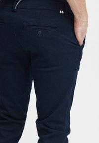 Marineblå chinos lavet af bomuld med en glat tekstur, der har baglommer med knaplukninger og et lille logo-patch i rød, hvid og blå.