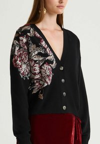 Cardigan noir boutonné avec un col en V profond et une broderie florale complexe sur l'épaule gauche, associé à une jupe en velours rouge profond.
