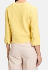 Žlutá texturovaná bunda s třičtvrtinovými rukávy, detailním prošíváním a bočními rozparky na lemu, spárovaná se světle zbarvenými kalhotami.