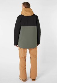 Bunt gefütterte Jacke mit schwarzem oberen Abschnitt, olivgrüner Mitte und beiger Kapuze, kombiniert mit beigen Schnee-Hosen mit Taschen und Reißverschlüssen.
