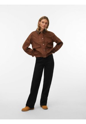 Femme debout, les mains sur les hanches, portant une veste marron, un pantalon noir et des chaussures beige sur fond blanc.