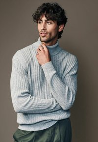 Hallhalli hall turtleneck-sweater pehmeast tekstuurist, millel on kõrge krae ja pikad varrukad, koos tumeroheliste pükstega.