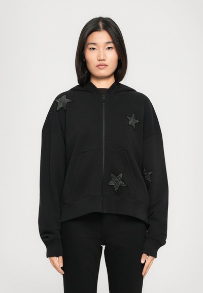 Woman wearing a black zip-up hoodie with four black star-shaped patches and black pants, standing against a plain light background.
