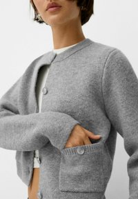 Grauer Wollcardigan mit rundem Ausschnitt, einfacher Knopfverschluss und zwei Fronttaschen. Weiche Textur und lockere Passform.