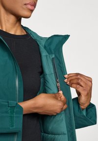 Teal pufferjack met een rits aan de voorkant en een zijzak. Gladde textuur, lichtgewicht materiaal, met kleurcontrasterende ritsdetails.