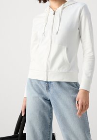 Sudadera blanca con cremallera, bolsillo canguro, cordones y puños acanalados, combinada con jeans azul claro y una bolsa tote negra.