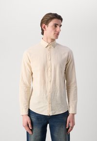 Light beige long-sleeve shirt with a button-down collar and front buttons. Soft texture, slightly wrinkled, paired with dark blue jeans.