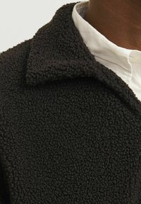 Schwarze, texturierte Fleecejacke mit Kragen, die eine weiche, flauschige Oberfläche aufweist. Darunter ist ein weißes Hemd mit Kragen teilweise sichtbar.