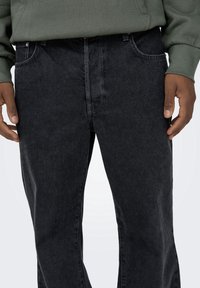 Jean en denim noir à coupe droite, avec des surpiq ûres contrastantes et des poches avant. Porté avec un sweat-shirt vert.